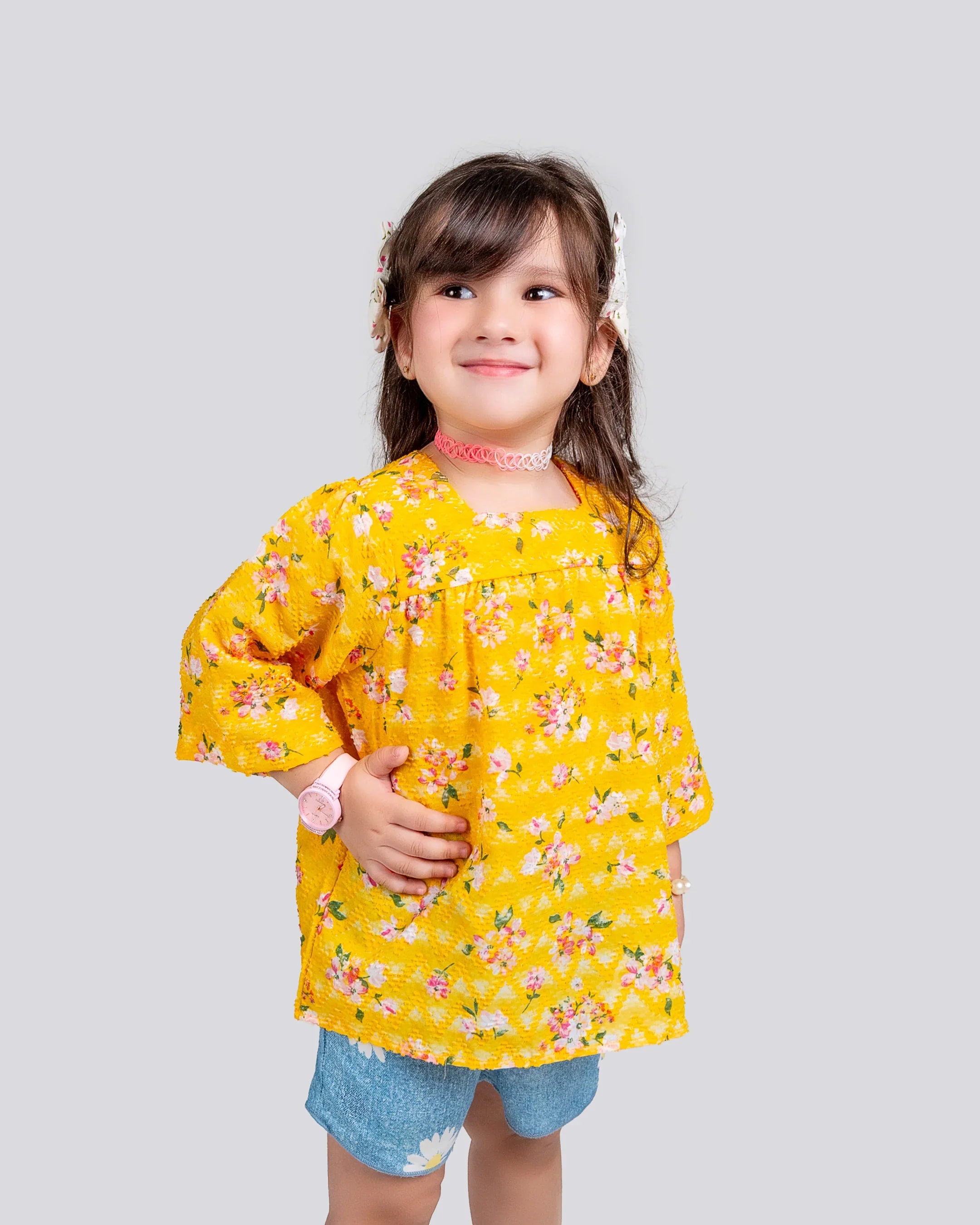 Young girl wearing a yellow floral blouse and blue shorts on a light gray background