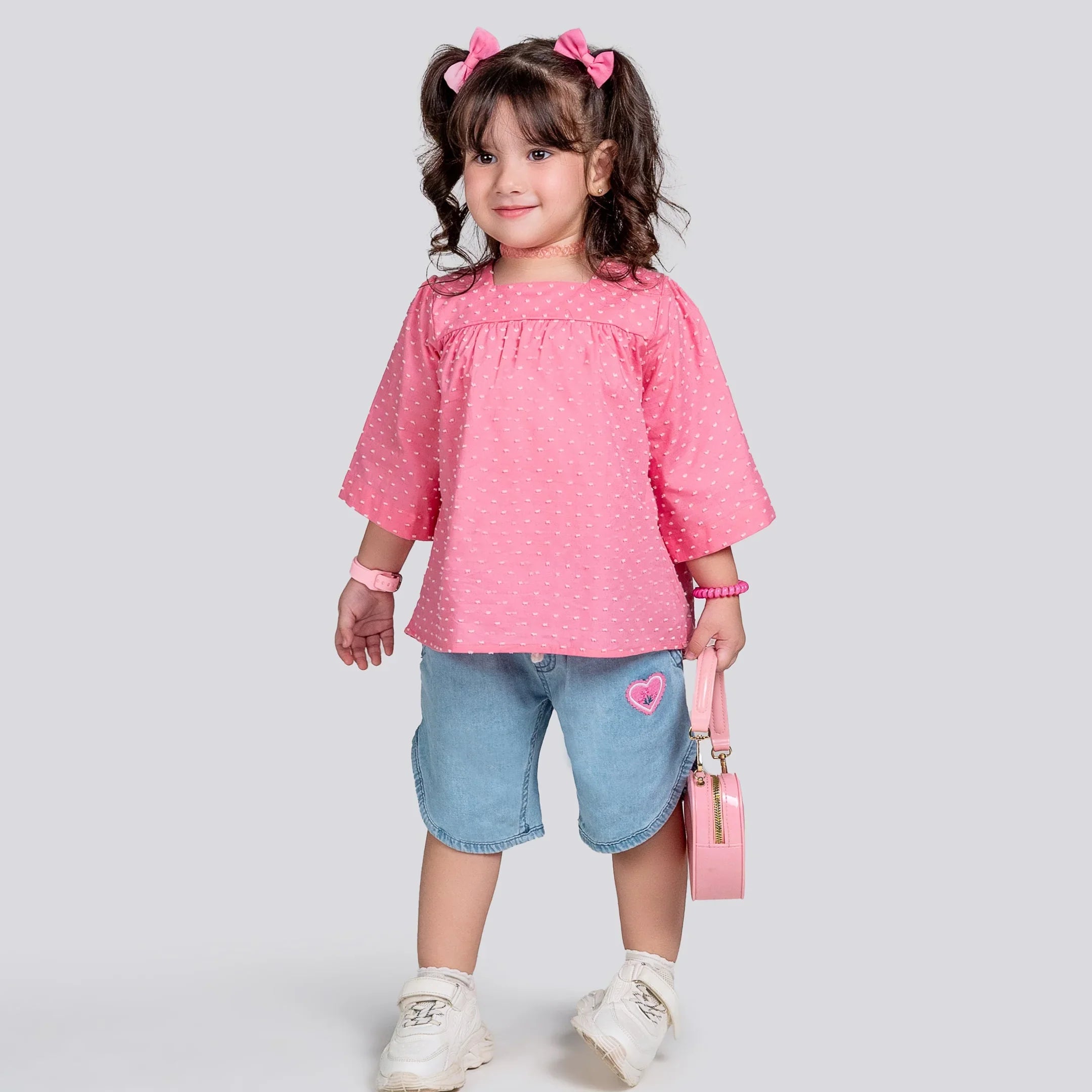 Child wearing a pink blouse and denim shorts with a heart design, holding a pink bag on a light gray background.