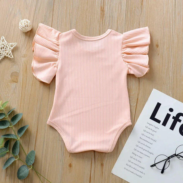 Pink baby romper with ruffled sleeves on a wooden surface with a magazine and glasses.