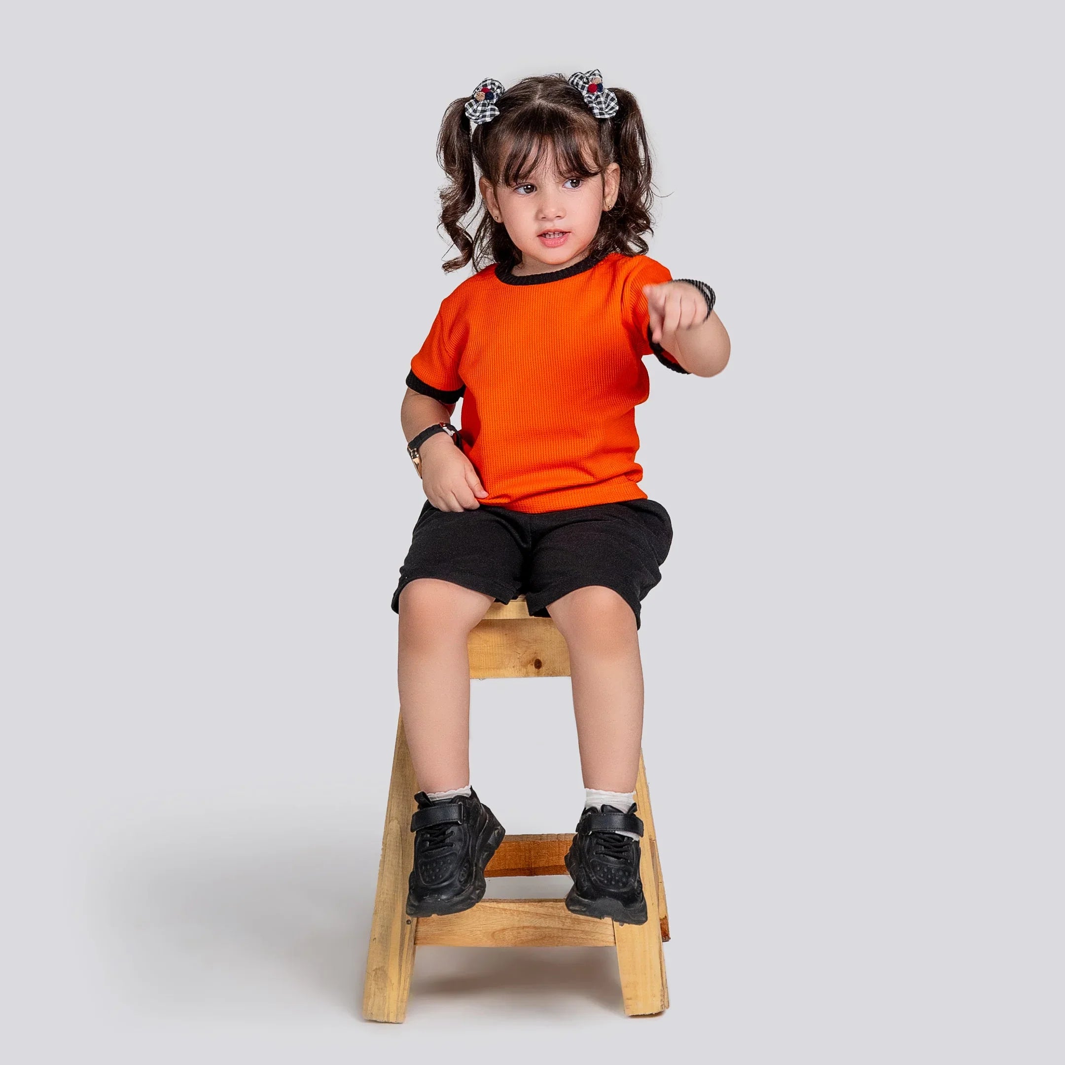Child sitting on a wooden stool wearing an orange shirt and black shorts against a plain background