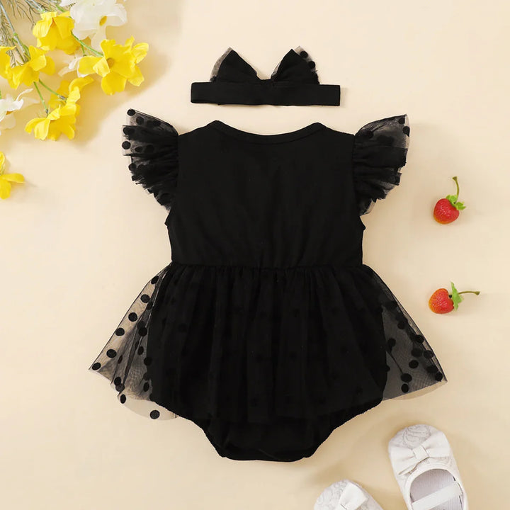 Black baby romper with tulle details and matching headband on a beige background with flowers and shoes.