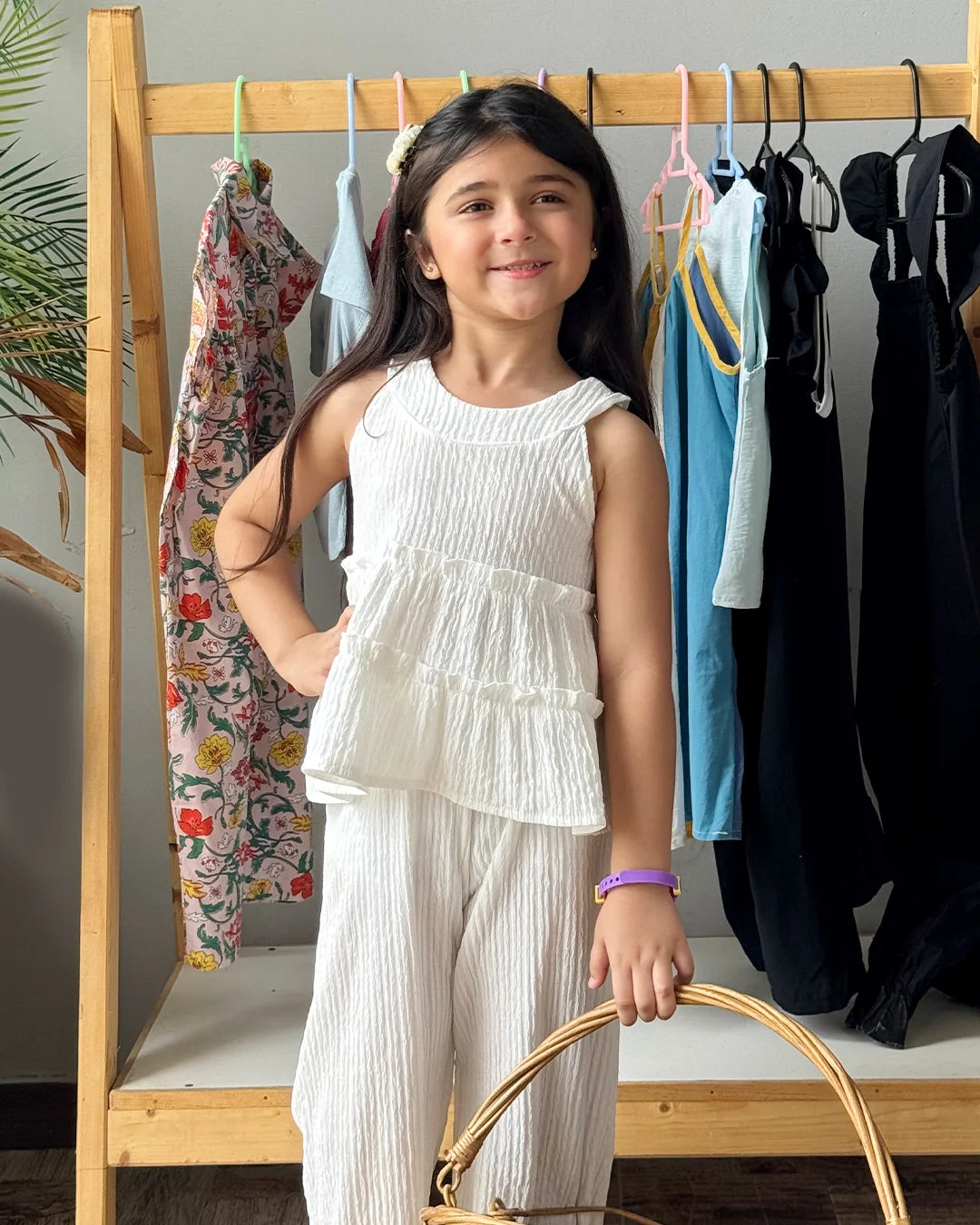 Young girl in a white outfit standing in front of a clothing rack with various garments.
