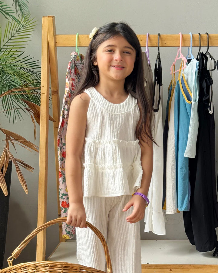 Young girl in a white outfit standing in front of a clothing rack with various garments.