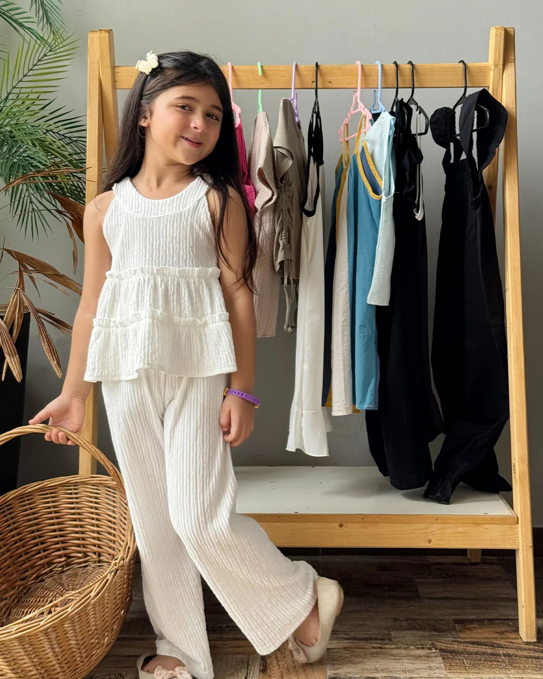 Young girl in a white outfit standing next to a wooden rack with clothes in a room.