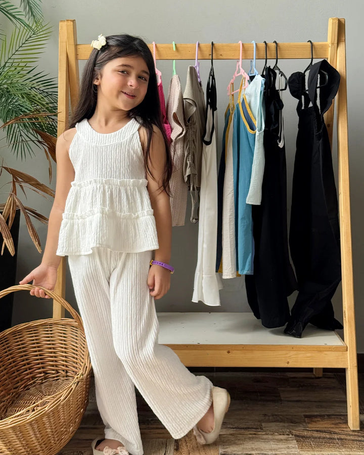 Young girl in a white outfit standing next to a wooden rack with clothes in a room.