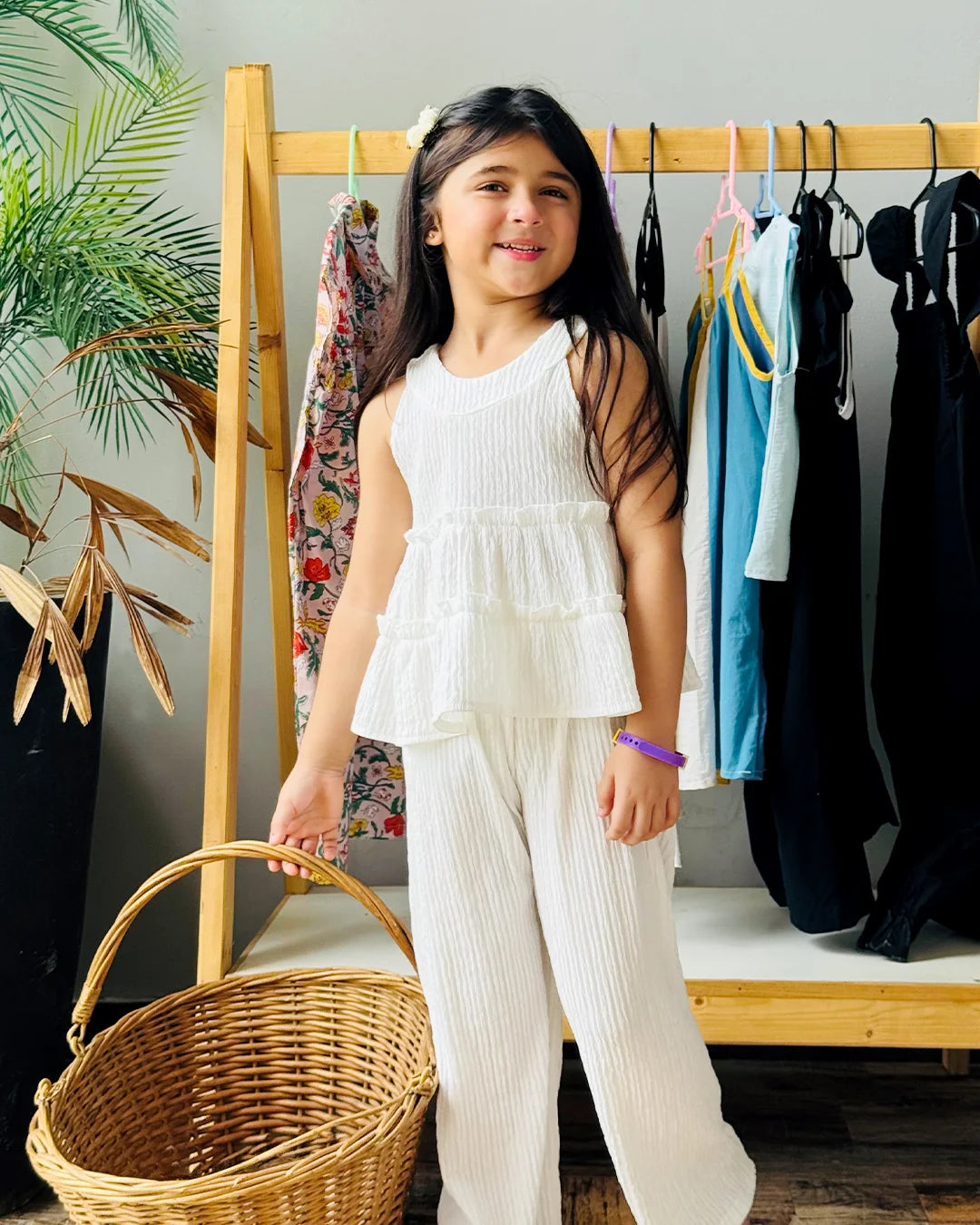 Young girl in a white outfit standing next to a clothing rack with a wicker basket.