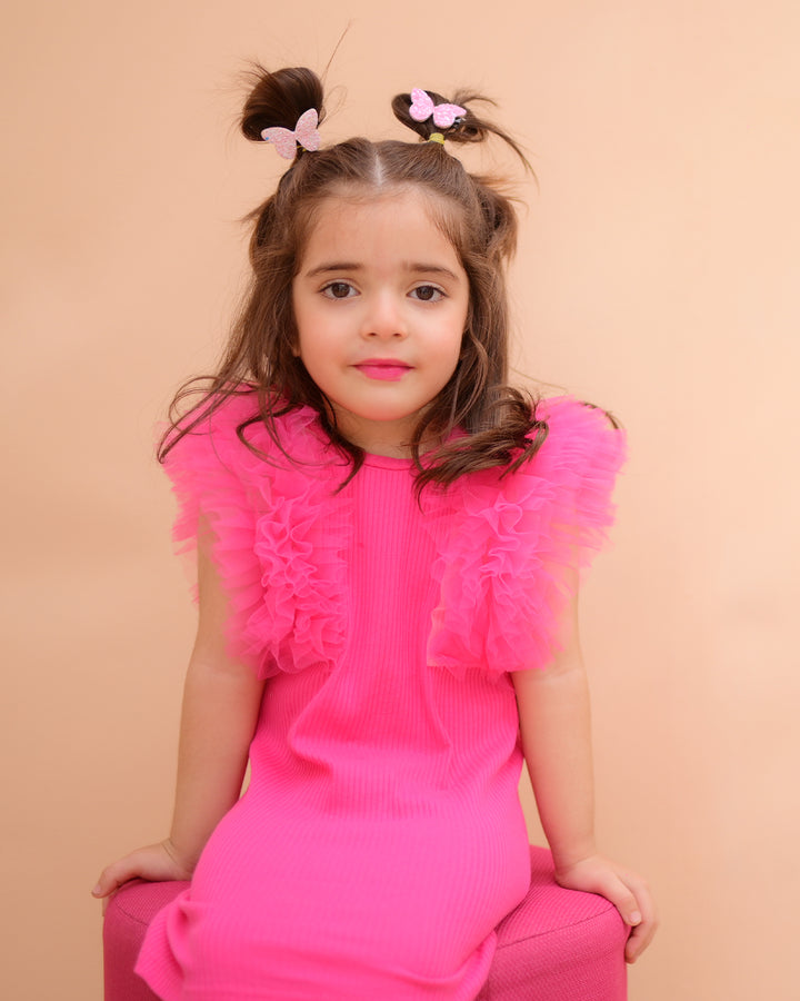 A girl wearing a pink sleeveless tulle dress with ruffle details on the shoulders.