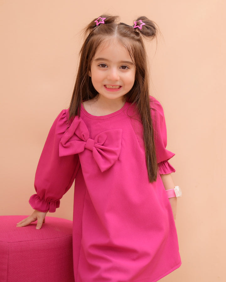 A young girl wearing a pink knee-high dress with long puff sleeves and a bow detail on the front, standing next to a purple cushion.