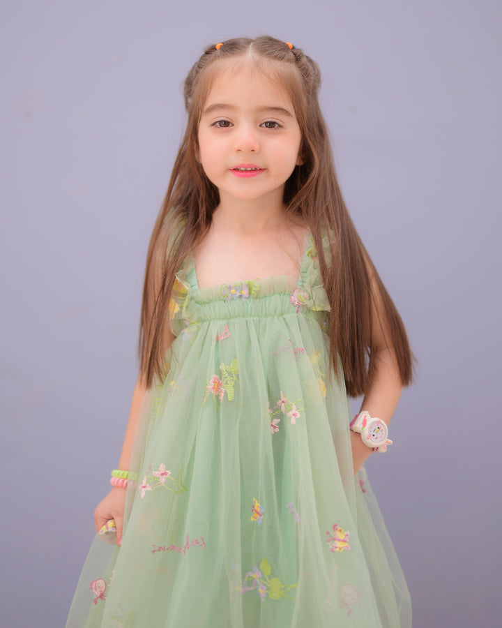 Young girl wearing a light green floral embroidered dress with ruffled straps, standing against a soft lavender background and smiling gently.