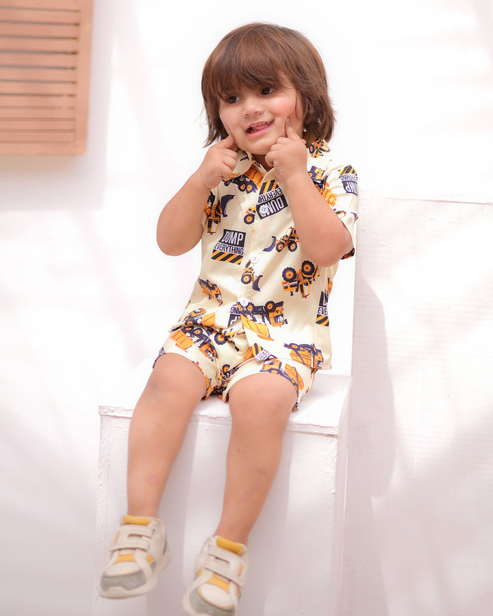A cheerful young boy sitting on a white ledge, smiling and pointing at his cheeks while wearing a yellow outfit printed with construction vehicles and white sneakers.