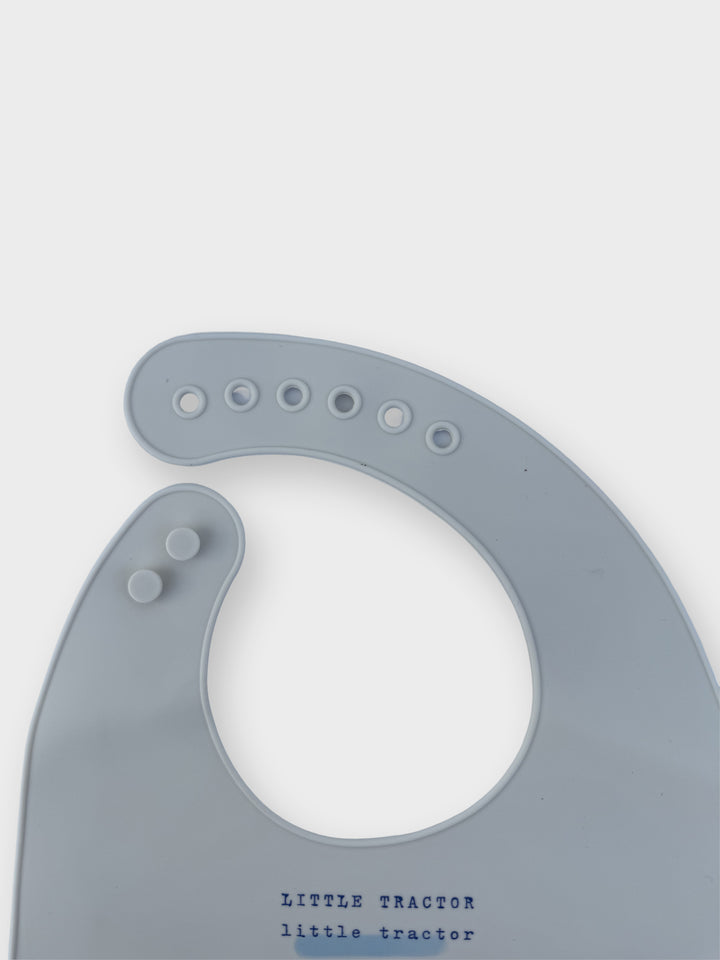 A light grey silicone baby bib with adjustable button fasteners and the text “LITTLE TRACTOR” printed near the bottom.
