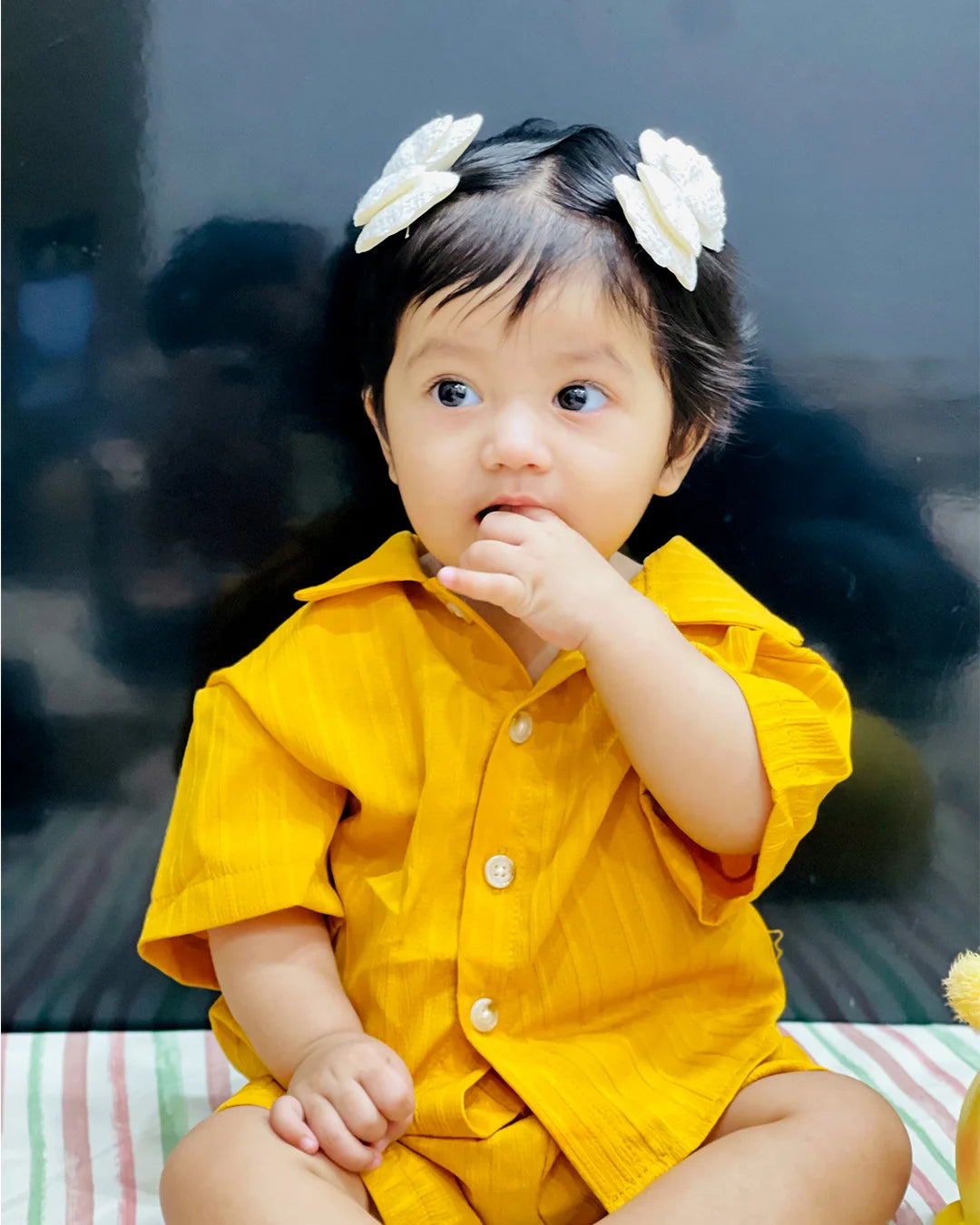 Child wearing a yellow outfit with white flower headbands sitting on a striped surface.