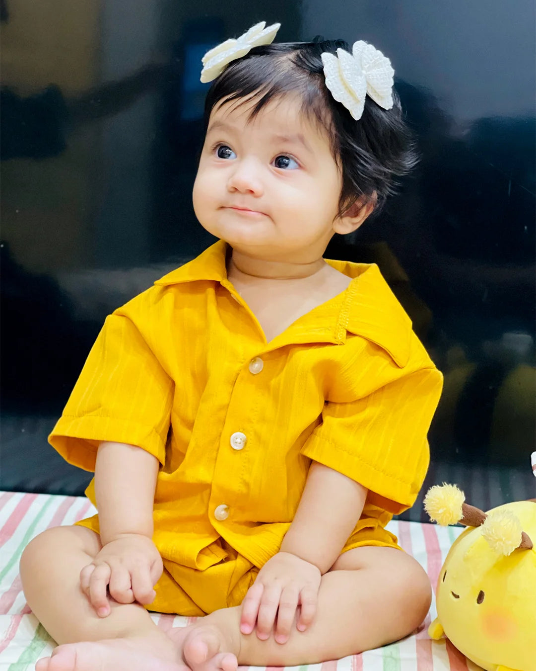 Child wearing a yellow outfit with white floral hair accessories, sitting on a striped blanket.