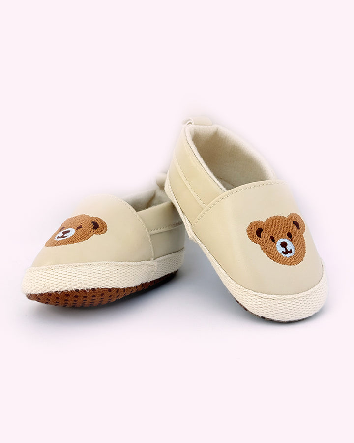 Pair of beige baby shoes with embroidered teddy bear faces on the front and soft rubber soles for grip, placed against a light pink background.