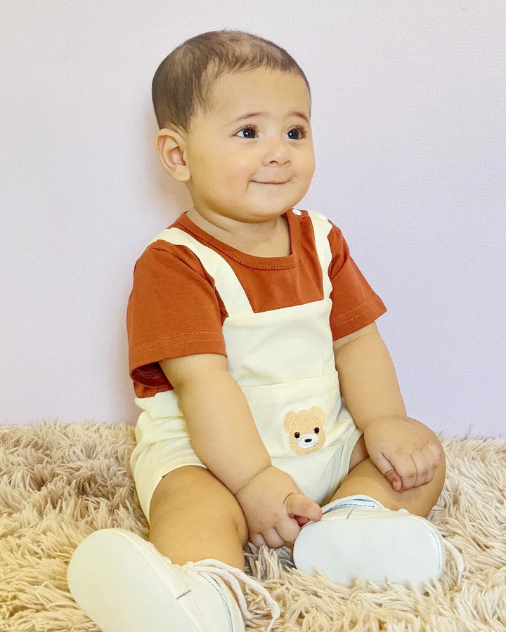 Baby wearing a red shirt and white overalls with a bear design, sitting on a beige rug.