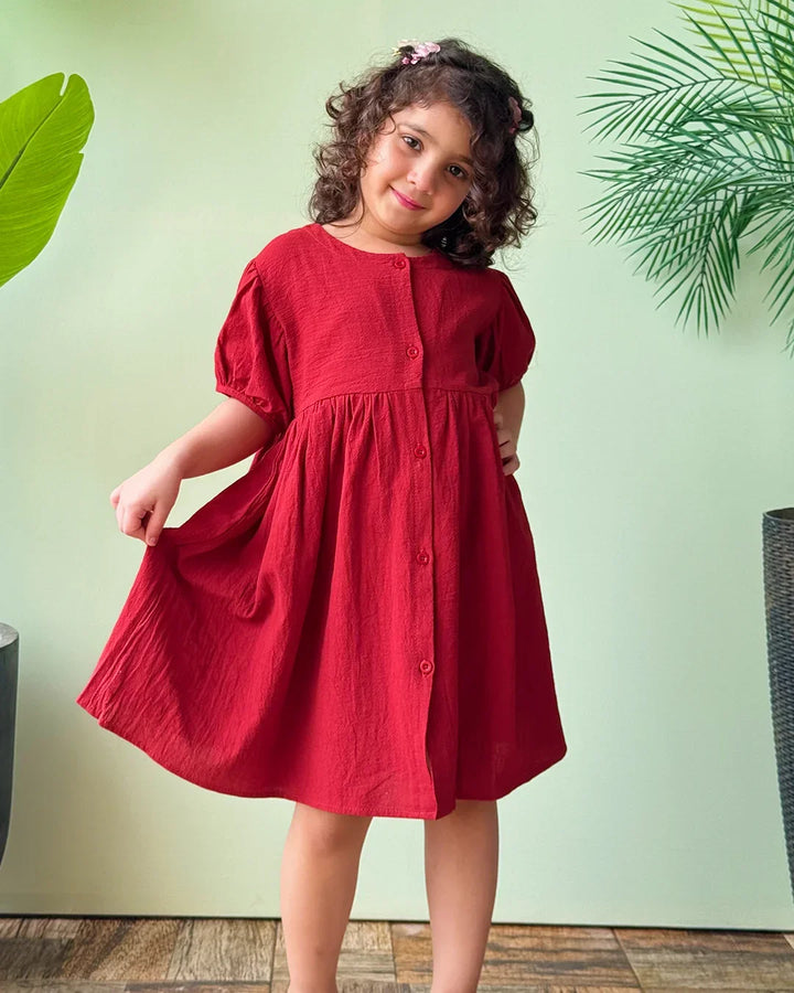Young girl wearing a red dress standing against a light green wall with plants.