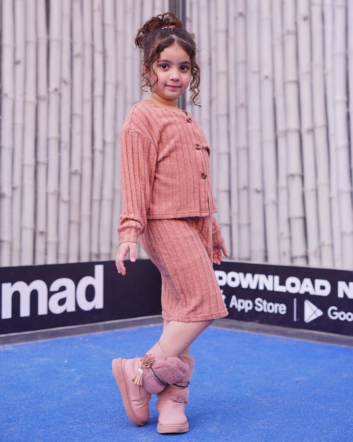 Child wearing a pink textured outfit with matching shoes on a blue surface with branding in the background.