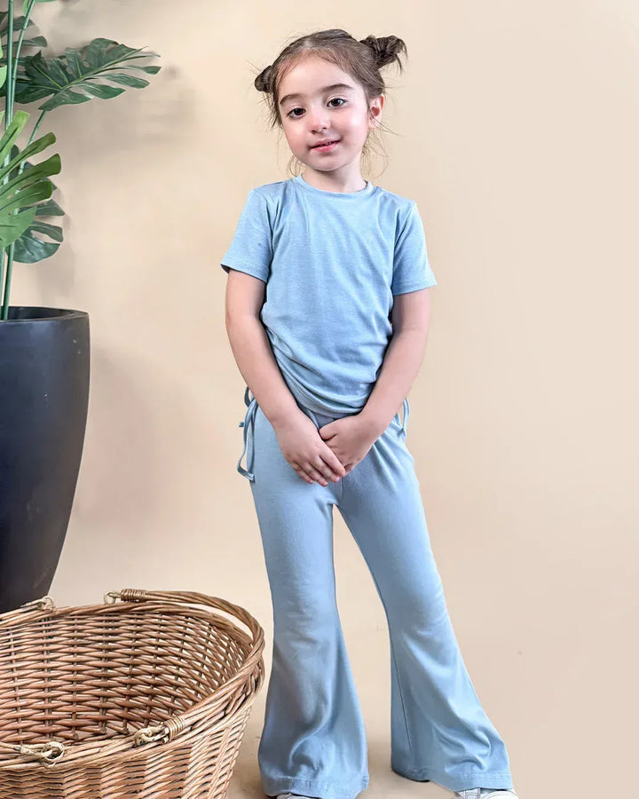 Young girl in light blue outfit standing next to a woven basket and plant against a beige wall.