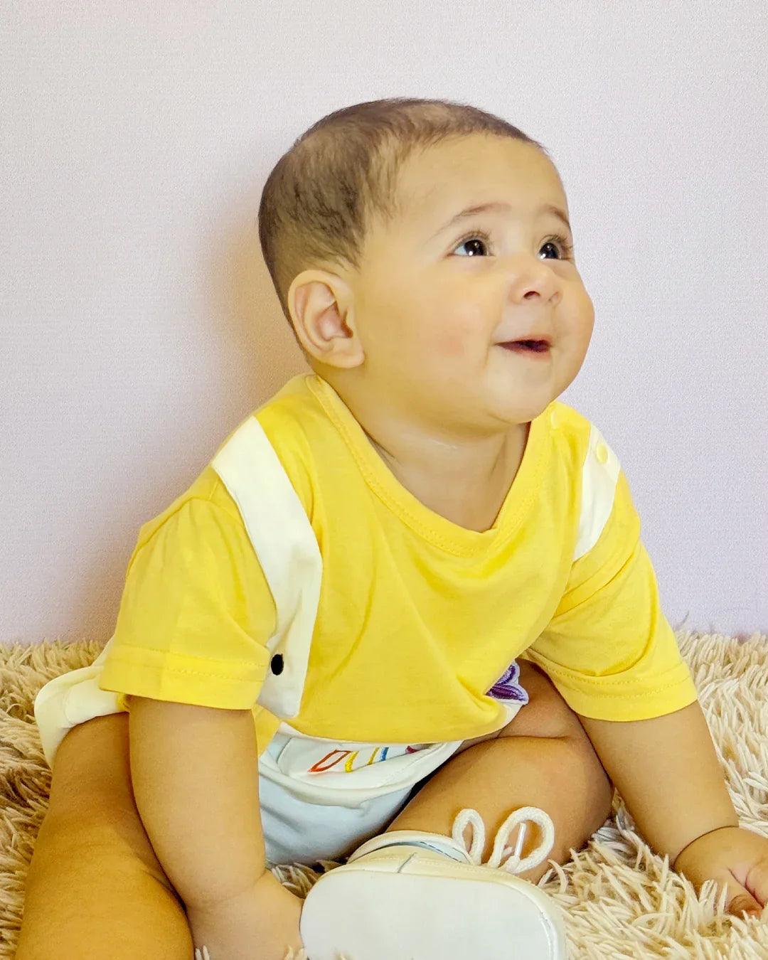 Baby wearing a yellow shirt sitting on a textured surface with a plain background