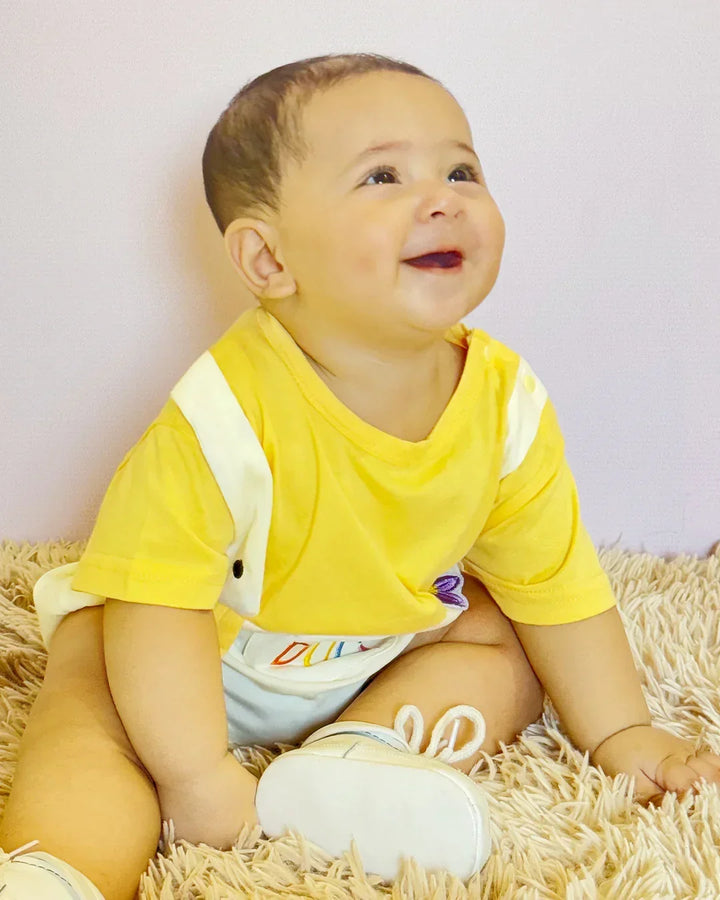 Baby sitting on a textured surface wearing a yellow shirt and white shoes.