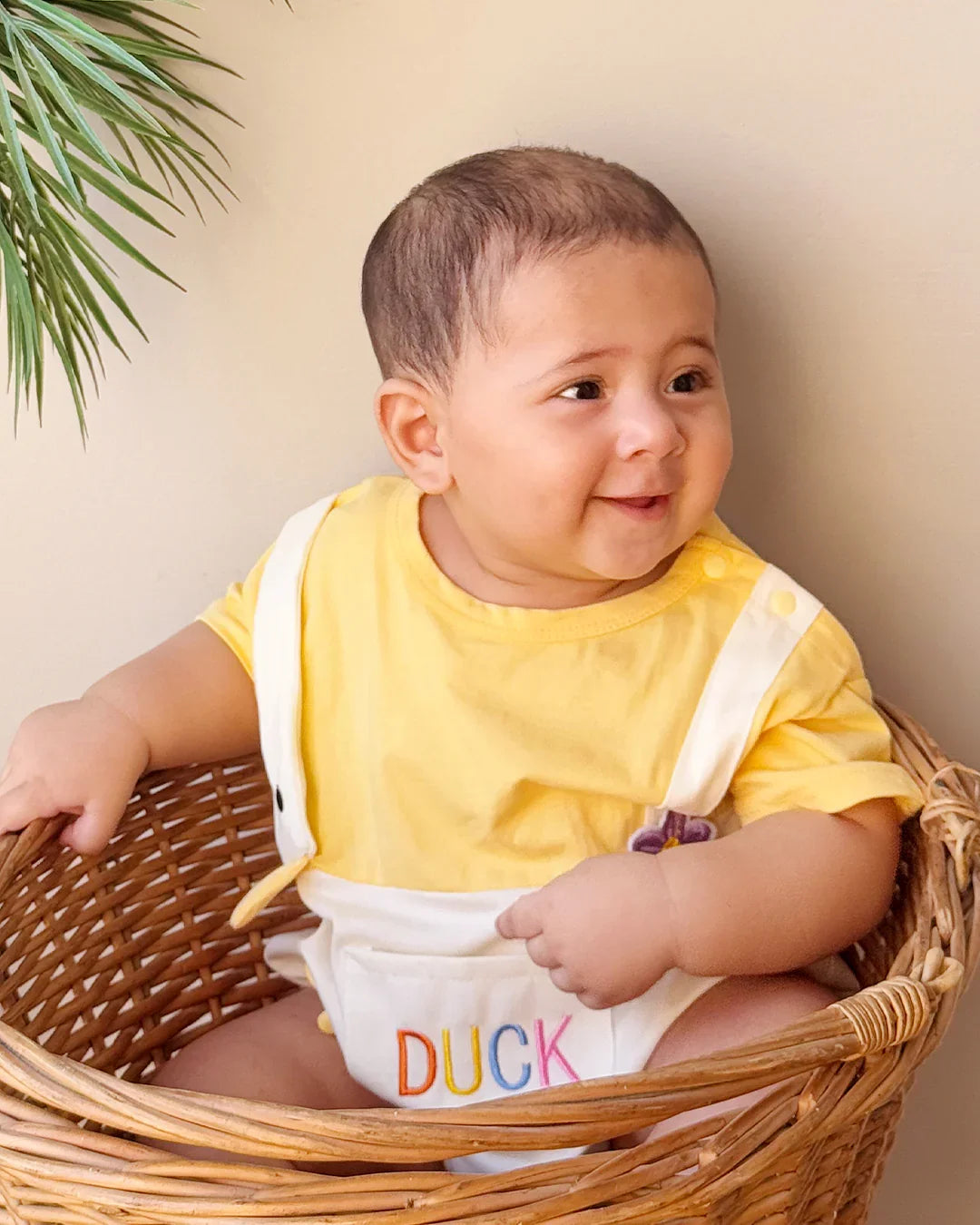 Baby wearing a yellow shirt and white diaper with 'DUCK' printed on it, sitting in a wicker basket.