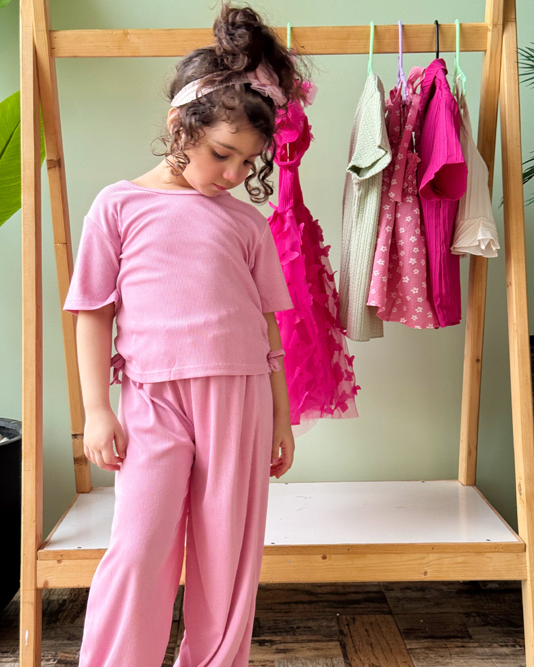 Child in pink outfit standing in front of a wooden rack with clothes