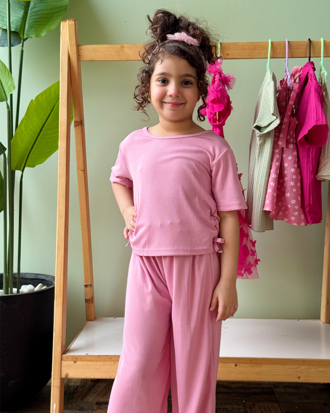 Young girl in pink outfit standing in front of a wooden rack with clothes.