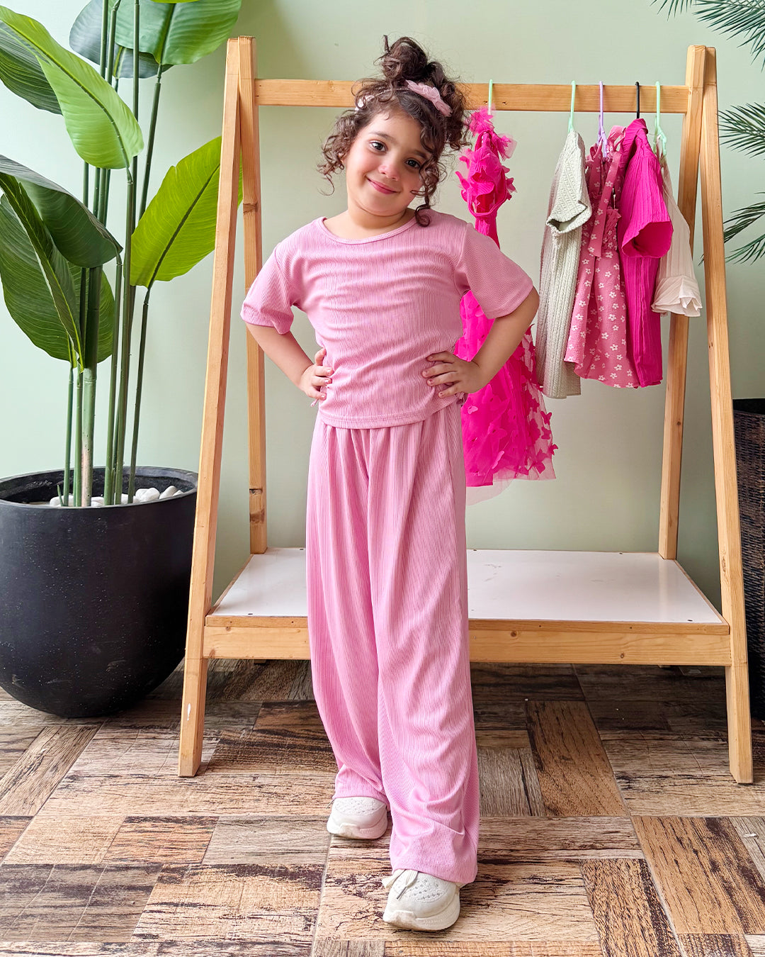 Young girl in a pink outfit standing in front of a wooden rack with clothes, surrounded by plants.