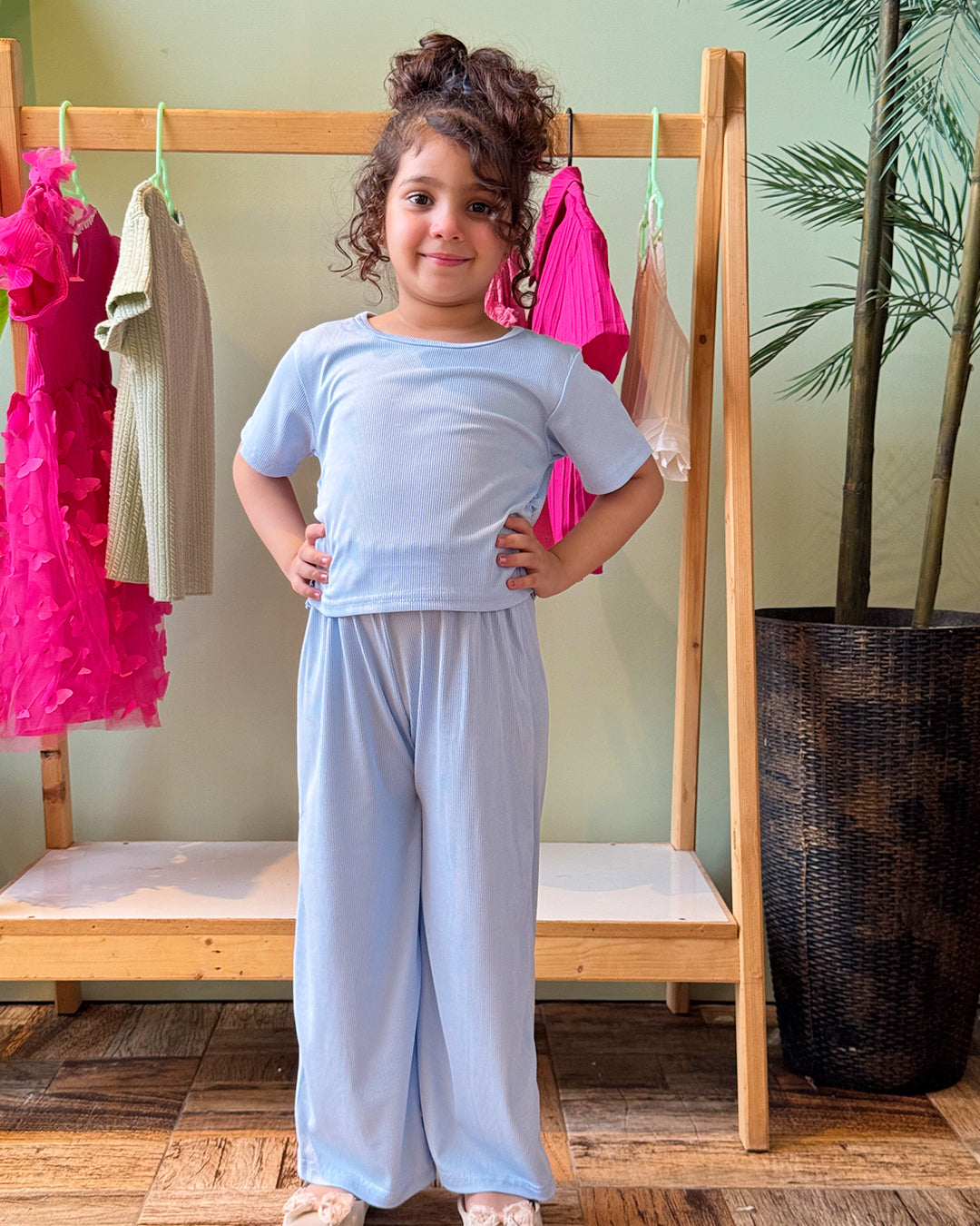 Child wearing a light blue outfit standing in front of a rack with pink dresses.