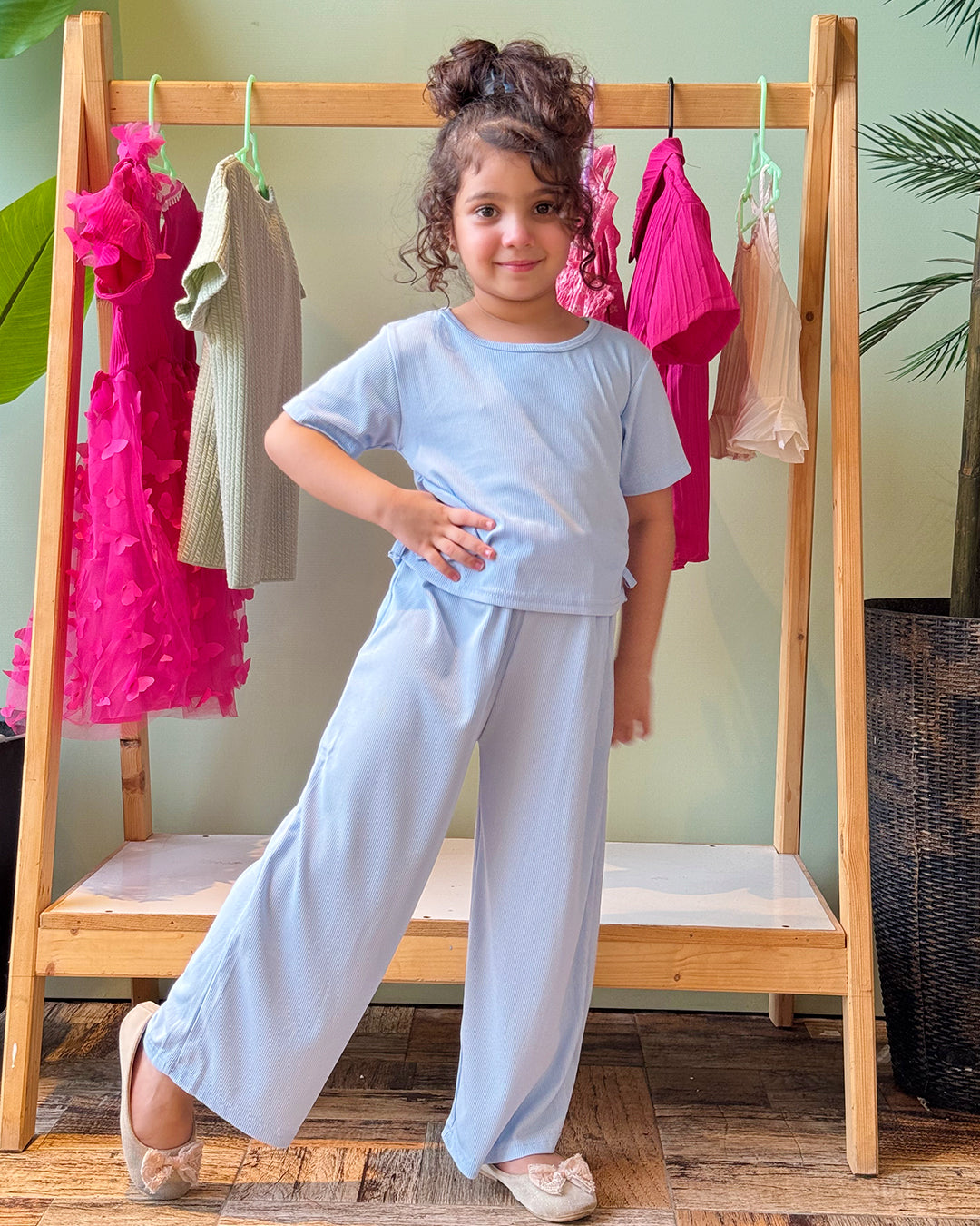 Young girl in light blue outfit standing in front of a wooden rack with colorful dresses.