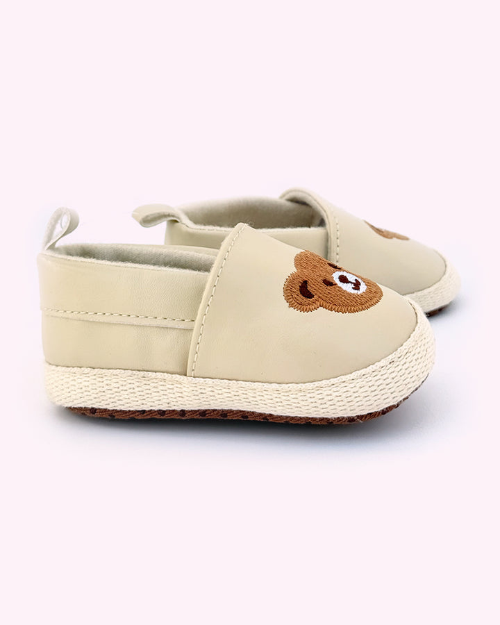 Pair of beige baby shoes with embroidered teddy bear faces on the front and soft rubber soles for grip, placed against a light pink background.