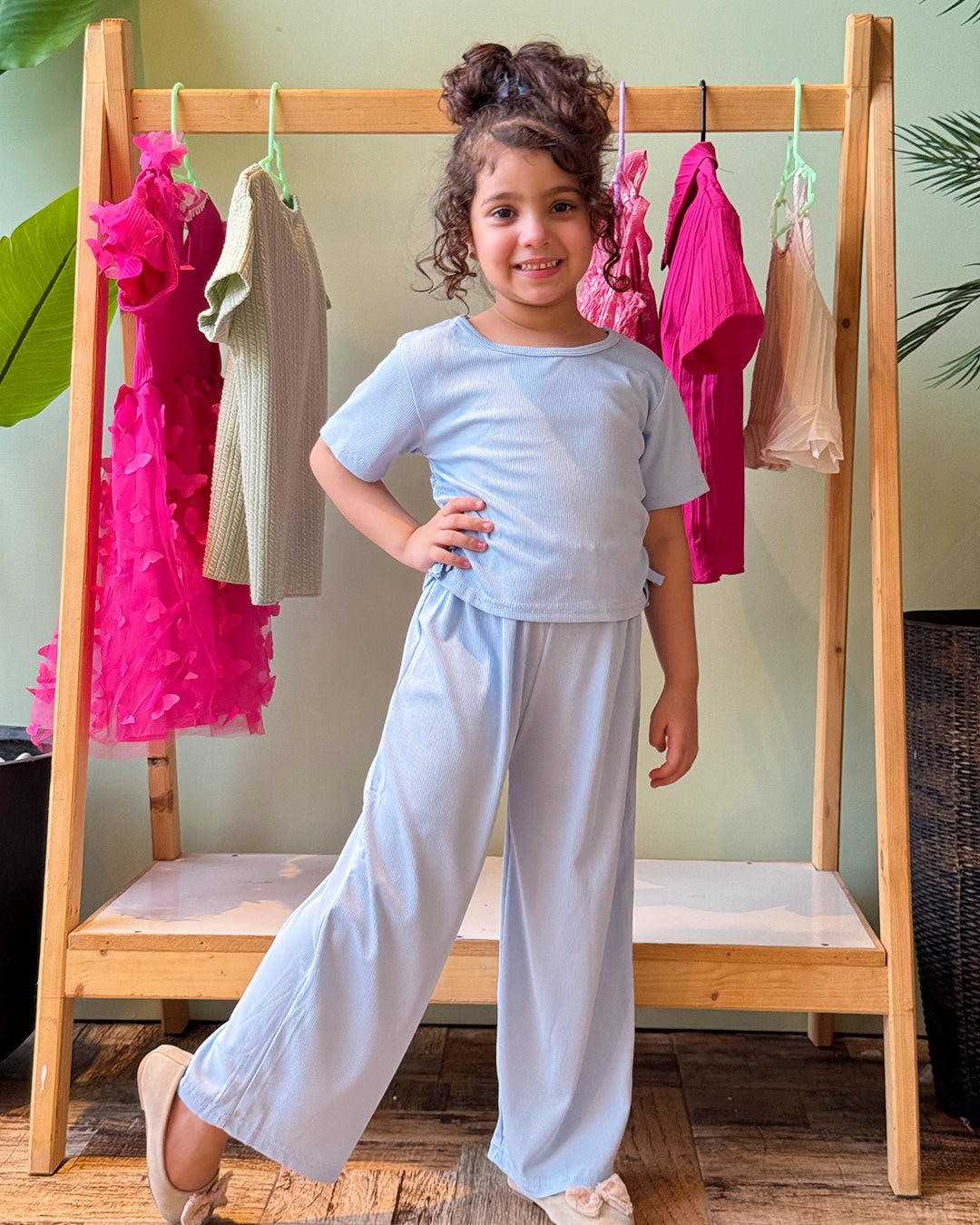 Young girl in a light blue outfit standing in front of a wooden rack with colorful dresses.
