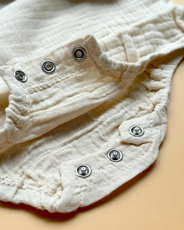 Close-up of a cream-colored baby onesie made from soft muslin fabric, featuring metal snap buttons for easy fastening and elastic edges for comfort.