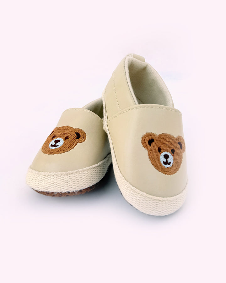Pair of beige baby shoes with embroidered teddy bear faces on the front and soft rubber soles for grip, placed against a light pink background.