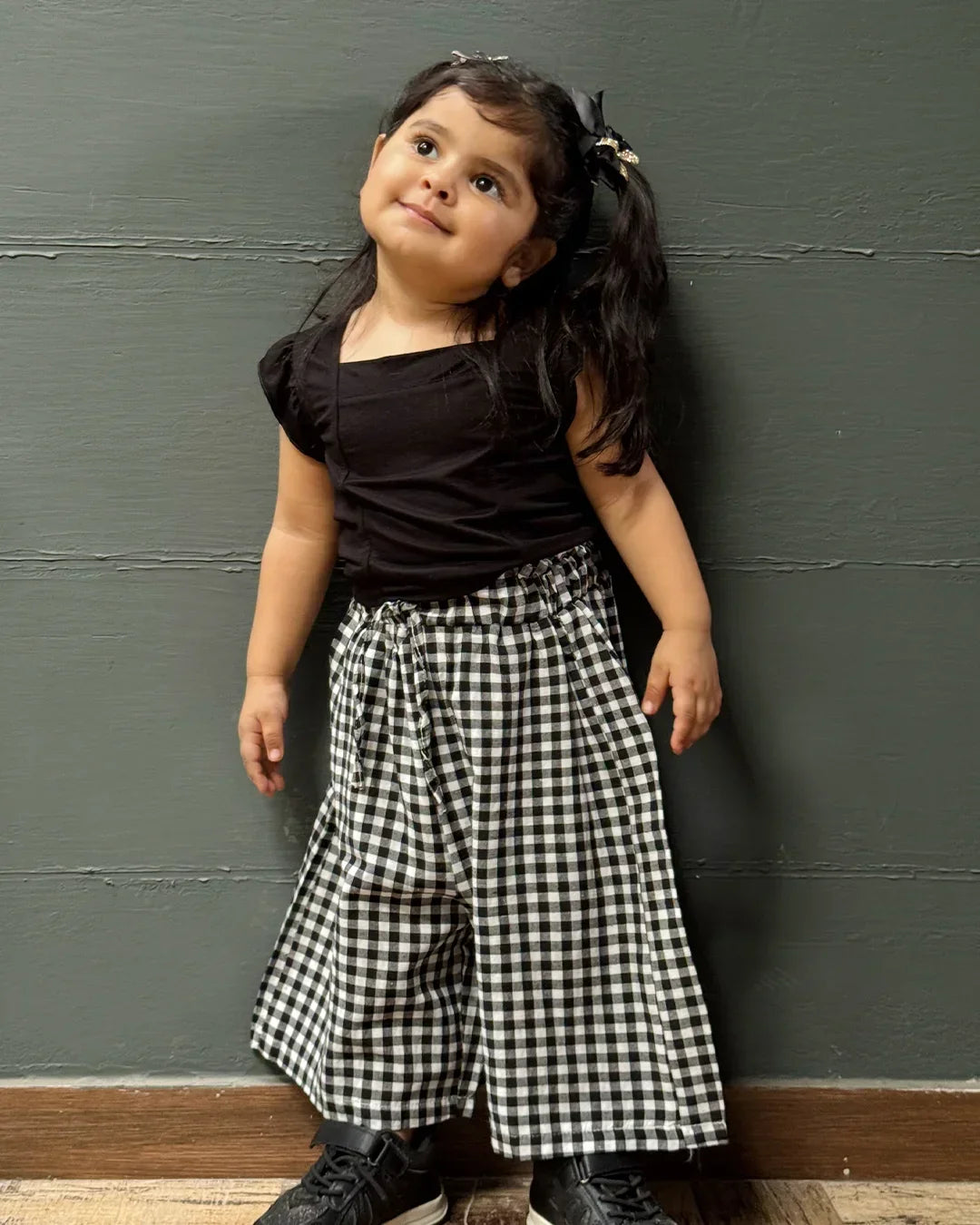 Child wearing a black top and black and white checkered pants against a dark wall.