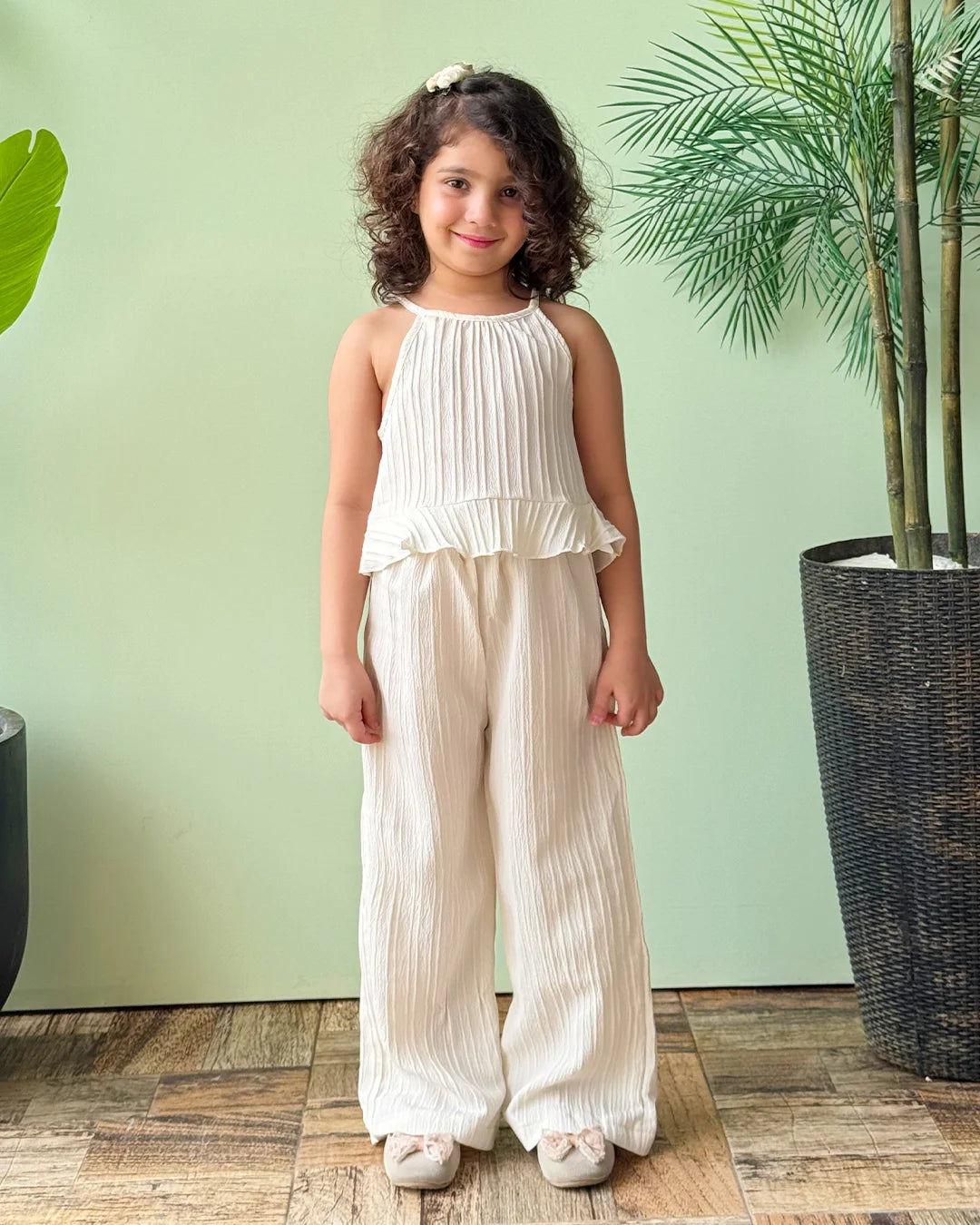 Young girl in a white pleated outfit standing in front of a green wall with plants.