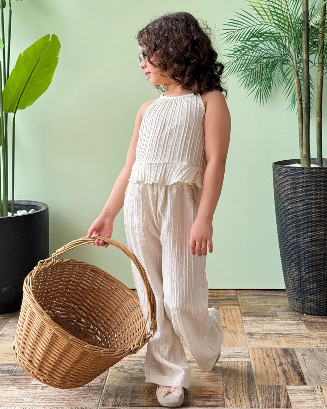 Child in a white outfit holding a woven basket indoors with plants in the background