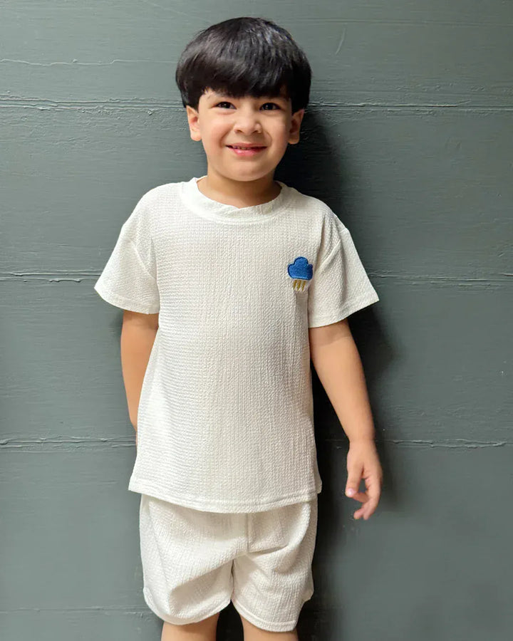 Child wearing a white t-shirt and shorts with a blue flower design against a gray wall.