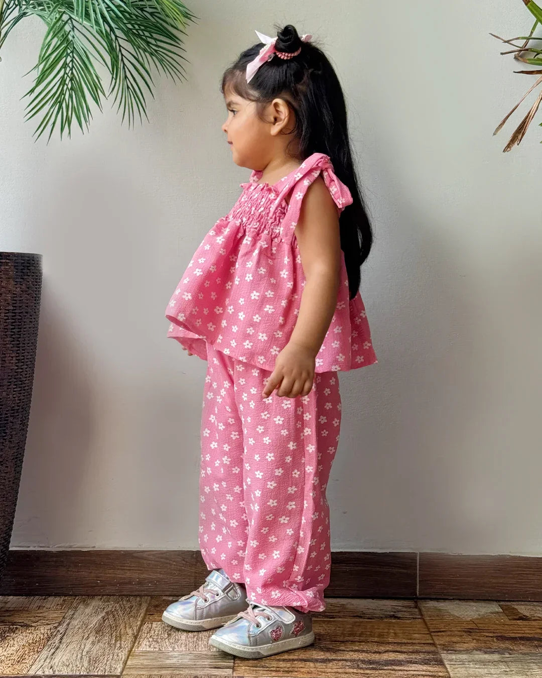Child wearing a pink floral outfit standing against a plain wall with plants in the background.