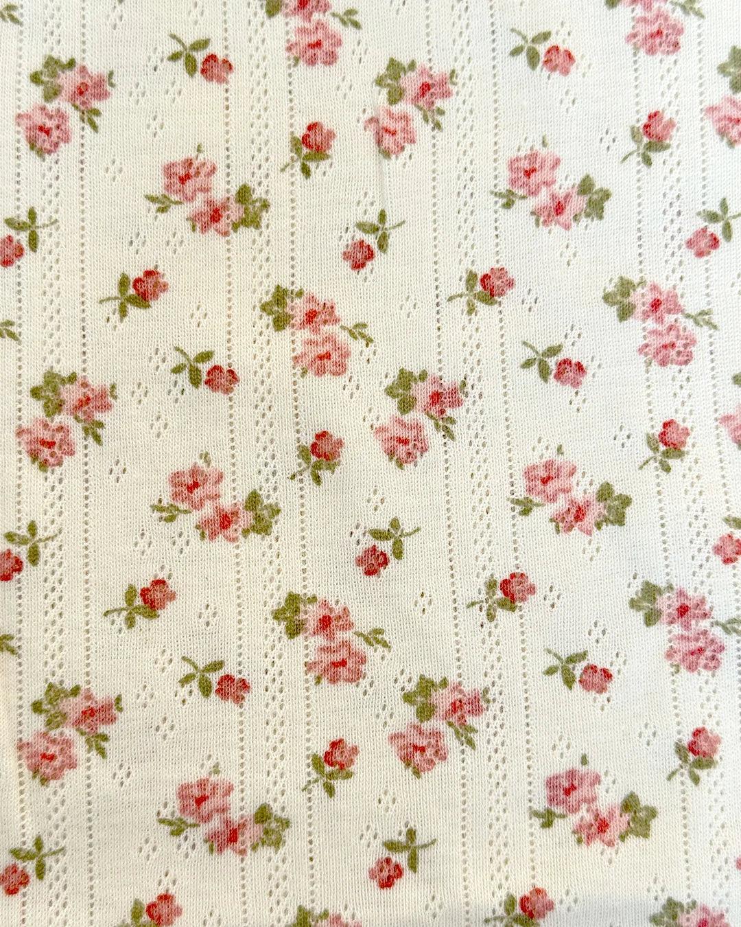 A soft cream-colored fabric with a delicate floral print featuring small pink roses and green leaves, arranged in vertical rows with subtle textured detailing.