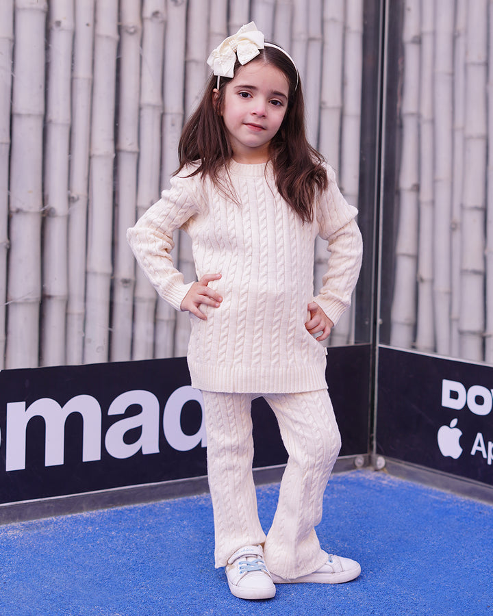 Young girl in a white cable knit outfit standing on a blue surface with a gray wall and Apple logo in the background.