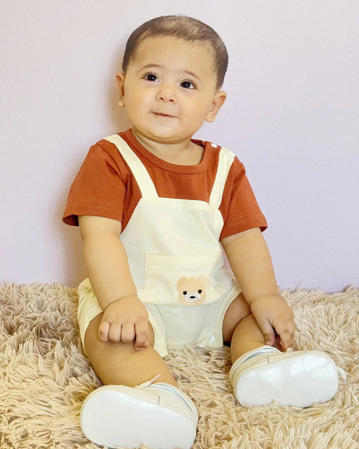 Baby wearing a brown shirt and white overalls with a teddy bear design, sitting on a beige carpet.