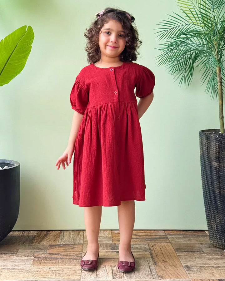 Young girl in a red dress standing in front of a green wall with plants.