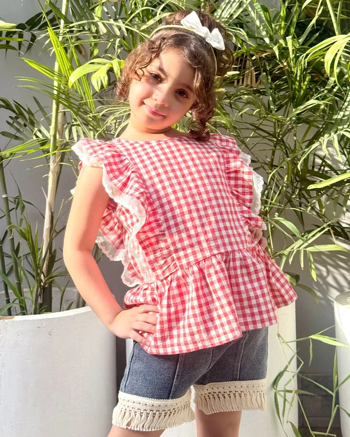 Young girl wearing a red and white checkered top with ruffles, standing outdoors with green plants in the background.
