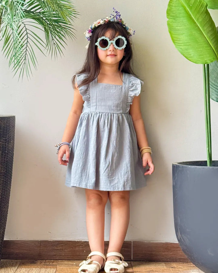 baby girl in a light blue dress with sunglasses and a flower crown, standing indoors with plants around.