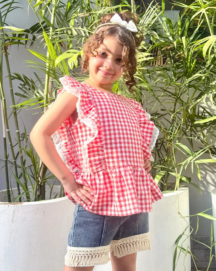 Young girl wearing a red and white checkered top with denim shorts standing in front of green plants.