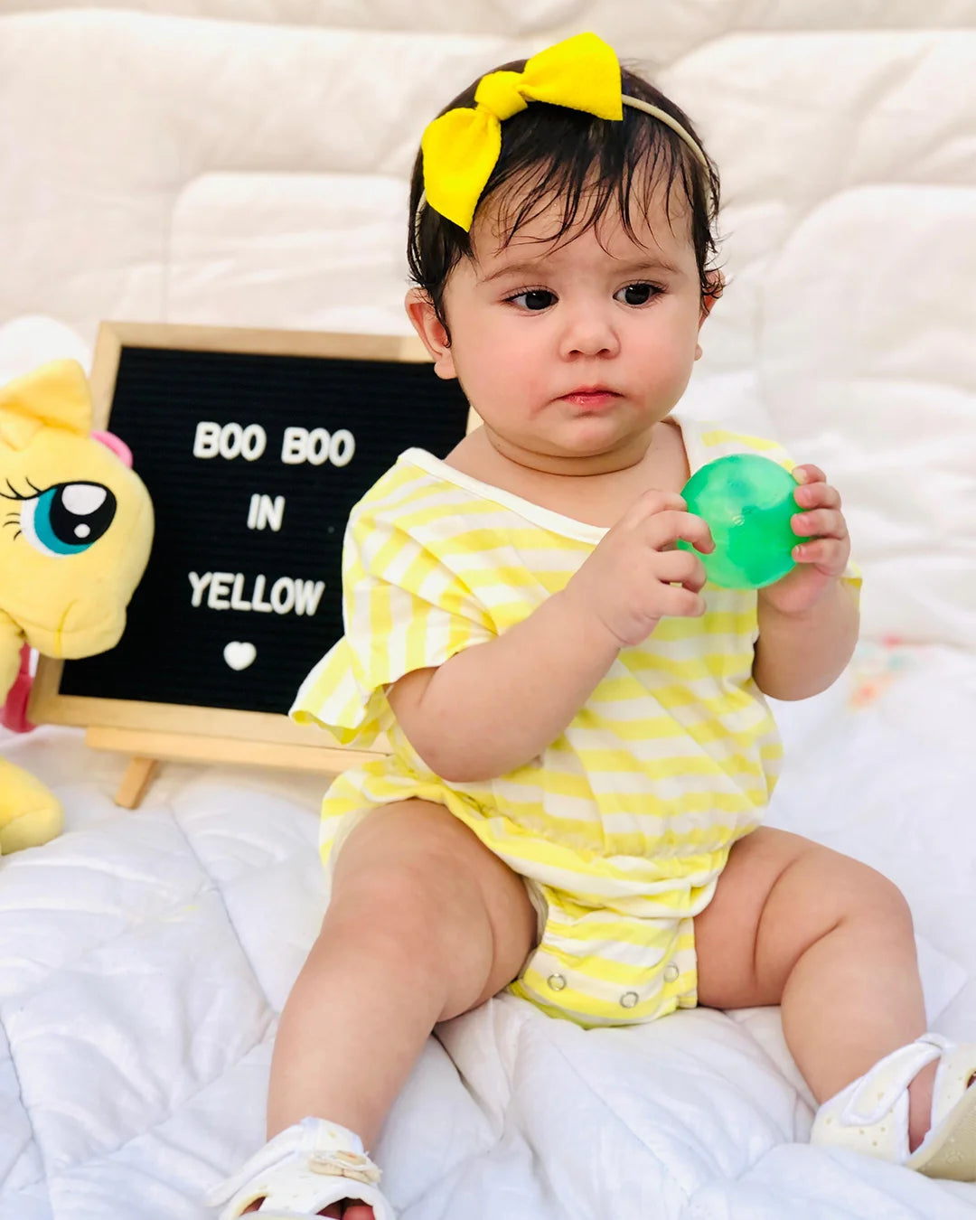 Baby in yellow outfit holding a green toy with a blackboard sign in the background