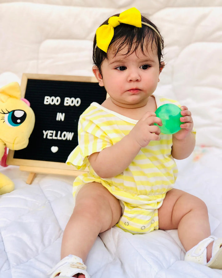 Baby in yellow outfit holding a green toy with a blackboard sign in the background