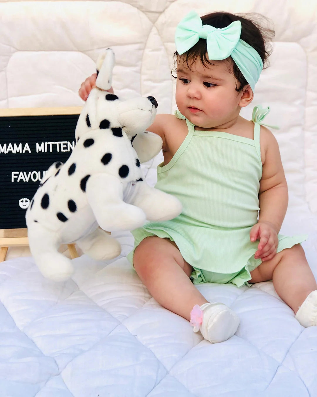 Baby in a green outfit holding a plush toy on a white blanket