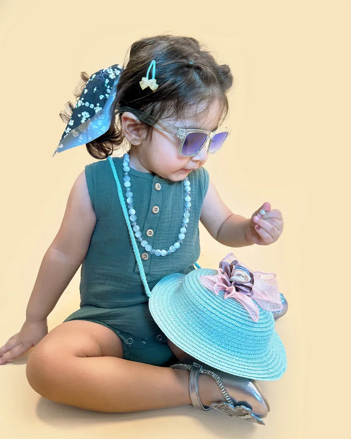 Child wearing a green outfit with sunglasses, holding a blue hat, on a beige background
